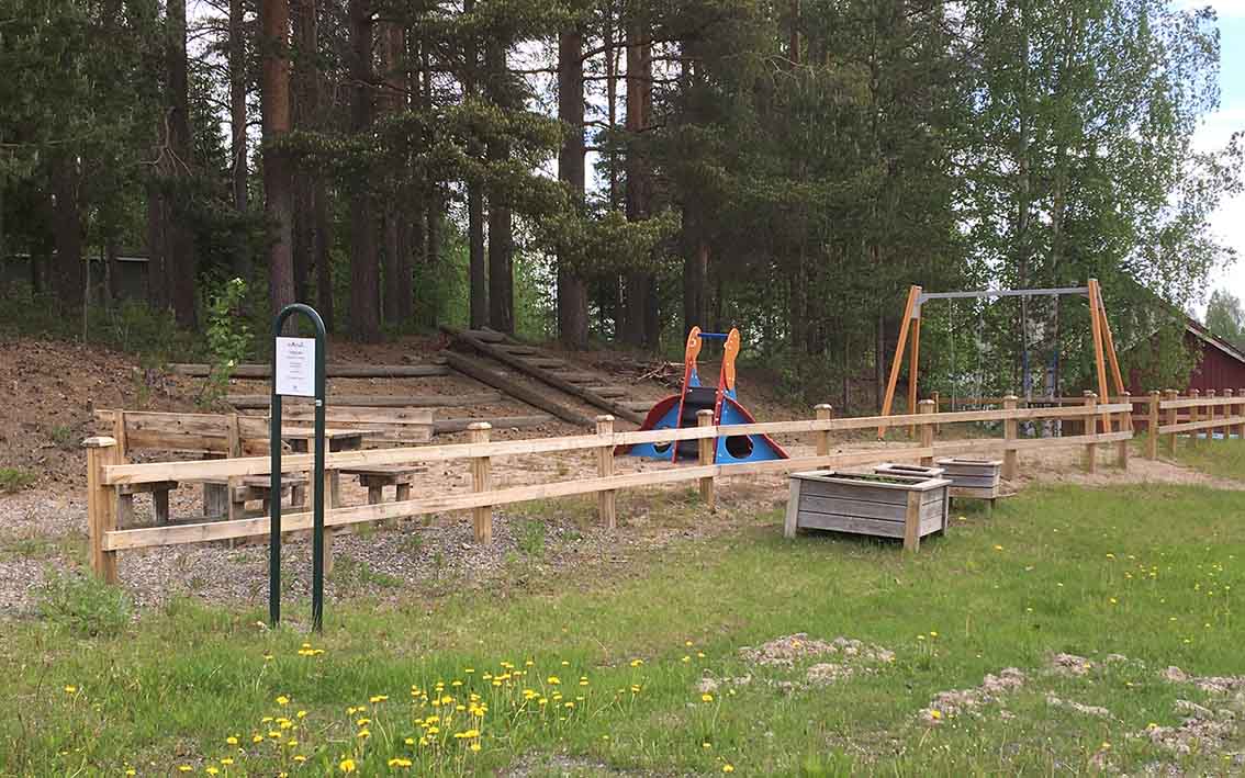 bord med sittplatser i kanten av lekplatsen. Ett lektorn i mitten och gungor längst bort. Upp mot skogen ligger stockar som det går att klättra i.