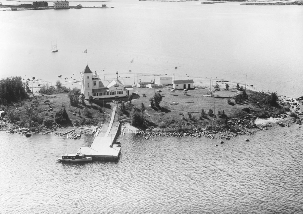 Flygfoto ner mot liten skärgårdsö med brygga och vitt trähus.