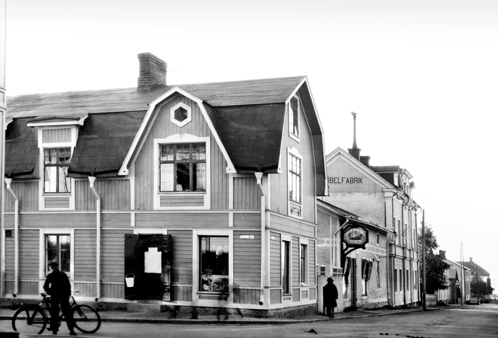 Trähus med butik i gatukorsning. Framför huset står en man med cykel.